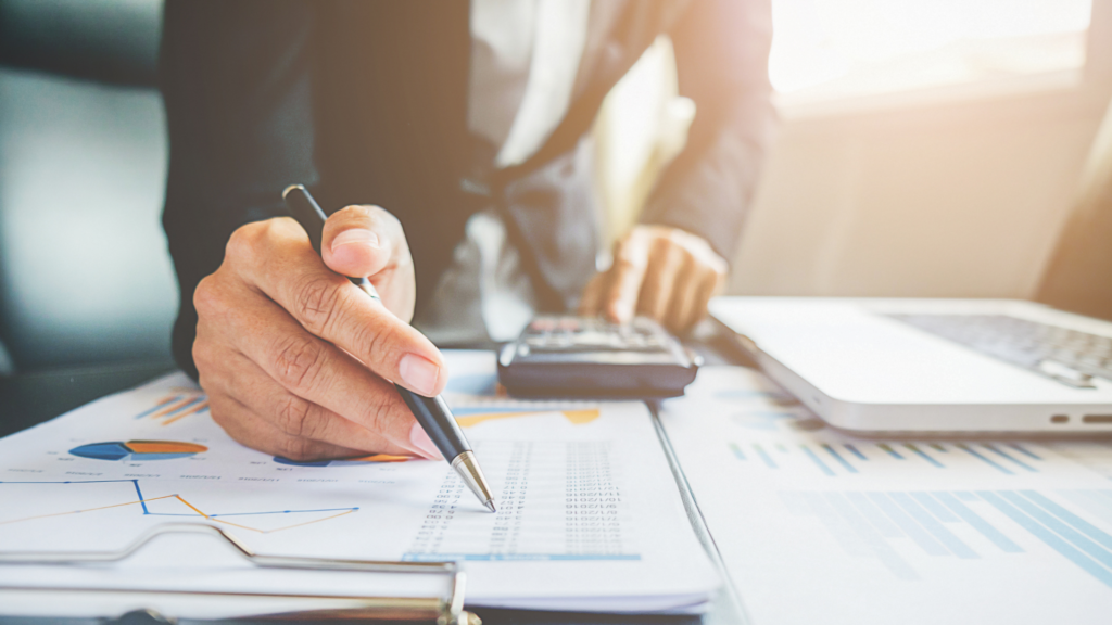 Contador analisando gráficos e dados financeiros enquanto faz anotações com uma caneta. Ao fundo, é possível ver uma calculadora e um computador, simbolizando a modernização das práticas contábeis. A cena reflete a preparação e a análise crítica necessárias para compreender e moldar o futuro da contabilidade.